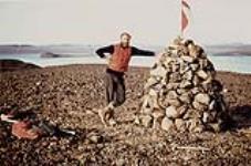 Man wearing a red vest standing next to a landmark of piled rocks. Arctic / Northern Canada [entre 17 juin-31 octibre, 1960].