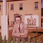 Un portrait informel du magistrat Jack White devant l'édifice du gouvernement à Woody Point, Bonne Bay, Terre-Neuve. Cet édifice comprend le bureau de poste, le bureau de la GRC, le bureau du magistrat ainsi que les ministères de la Foresterie et du Bien-être. August 1960