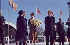 Princess Elizabeth holds flowers on stage, Prince Philip and several people stand nearby. Royal Visit 1951, Ontario. [Between October 8th and November 10th 1951].