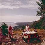 [A man holding a fish next to a woman standing by a picnic table with some seated children overlooking the water, Nova Scotia] août 1956