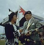 Betty Chan, a Chinese Canadian, admiring Pipe Major Bill MacLeod's tie, made from the MacLeod tartan at the festival in Winnipeg's Kildonan Park. Winnipeg, Manitoba.  1960