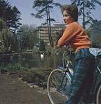 An attractive young cyclist pausing by the lagoon on her tour through beautiful Beacon Hill Park, Victoria, B.C. 1961