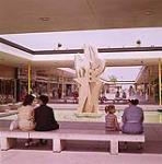 The shopping mall at Polo Park shopping centre, Winnipeg, Manitoba. A contemporary sculpture can be seen and four people on a stone bench.  [Le centre commercial du parc Polo, Winnipeg, Manitoba.] juin 1961