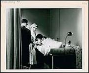Infirmière surveillant un patient allongé sur un lit d'hôpital. Un homme vêtu d'un complet observe. Infirmières et soins. Ministère de la Citoyenneté et de l'Immigration, division de l'information [between 1930-1960]