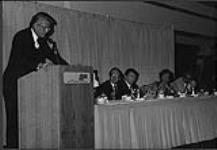 Tommy Hunter speaking at a podium at the Edmonton Convention Centre, seated at the head table are Larry Donohue (CFCW), Paul Mascioli (President, CCMA), Jo Walker-Meador (CMA), Bill Maxim (AACE) [between 1990-2000].