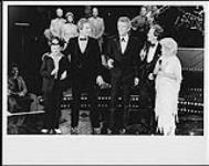 Tommy Hunter (centre), Pat Boone (?), Little Jimmy Dickens, and Carroll Baker (?) standing on a stage with two men, with a band at the rear of the stage [ca. 1982].