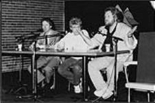 Trois hommes assis à une table, peut-être en train d'être interviewés. Il s'agit peut-être Randy Bachman de Bachman-Turner Overdrive (BTO). Edmonton [between 1973-1977].