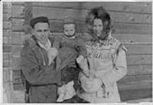 Man holding child, standing with woman wearing parka outside the old mission hospital, Fort Simpson, Northwest Territories juillet 1975