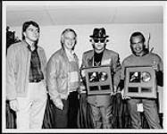 Elton John and Bernie Taupin receiving a Platinum album award for "Sleeping With The Past," at Toronto's Skydome - (l to r) Randy Lennox (VP Sales, MCA Canada), Ross Reynolds (Executive VP and GM, MCA Records Canada), Elton John, Bernie Taupin [ca. 1989].