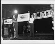 Shania Twain et un homme non identifié tenant une peinture d'un orignal, sur scène lors d'un événement télévisé de la musique country (CMT) [between 1995-1997].