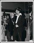 Ronnie Hawkins singing into a microphone onstage, with band members [between 1970-1975].
