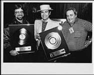 Elton John receiving platinum awards after a concert in Toronto: (l to r) Garry Newman (WEA VP Sales), Elton John, Roger Desjardins (WEA Artist Relations Manager) [ca. 1982].
