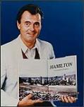 Unidentified man wearing a sports jacket and holding open a book entitled "Hamilton: A Community in Symphony" [ca 1986].