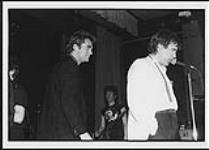 Huey Lewis on stage with an unidentified man at the microphone [between 1984-1986]