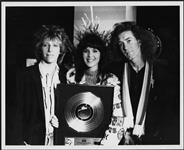 An unidentified woman, holding a Gold Record, is accompanied by two men [entre 1985-1990].