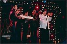 Four rock and roll impersonators holding a cake, while Bobby Curtola applauds a member of the audience [entre 1988-1995].