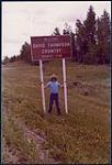 David Thompson holding on to a road sign with David Thompson Country Tourist Zone written across [entre 1978-1987].
