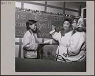 Leah Idlout holding an open book and facing two other Inuit girls. They are standing in front of a chalkboard with the names of colours in English, Resolute Bay (Qausuittuq), Nunavut September 1957.