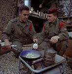 Sergeant Pearce right waits patiently whit Corporal Ryan cooks up a pan of bacon and a pot of coffee during pause for 3 MECH Cdo during Ex Refroger '74 in Bavaria 1974.