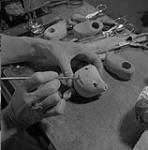 Mme Betsy Howard peignant le visage d'une poupée avec du vernis dans son atelier. On aperçoit des accessoires et des outils sur la machine à coudre juillet 1956