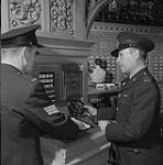 Prises de vue de l'ONF de la Bibliothèque du Parlement - le sergent F.G. Angrignon tenant une horloge de pointage et le constable H. Mongeon au tableau de contrôle de l'alarme d'incendie April 1957.