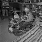 A.J. Jackson in his studio at Manotick, Ont. Here he is showing his grand-nephews and grand-niece the snowshoes he wore on Laurentian sketching trips which earned his habitant nickname Père Racquette - Father Snowshoes April 1959.