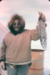 Man wearing a cloth parka trimmed with fur holding an Arctic Char, at Ekalluktuk River, Victoria Island 1962.