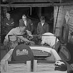 Stock Car Story. While friends look on, two members of the Winnipeg Roadsters Stock Car Racing Club are working on racing car motor. Crash helmets are a must in any motor sport September 1959.