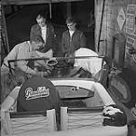 Stock Car Story. While friends look on, two members of the Winnipeg Roadsters Stock Car Racing Club work on racing car motor. Crash helmets are a must in any motor sport September 1959.