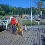 [Three men walking on a wooden dock near a boat] [between 1900-1976]