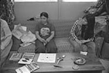 [Kenojuak Ashevak sitting on the couch at home as daughter Pee Ashevak looks on] December 1980