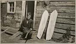 [Anishinaabe man sharpening axe at entrance of wooden house. Snowshoe boards are leaning against the wall to his right] 1920