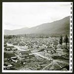 Lemon Creek with its orderly rows of small houses and patchwork-quilt gardens lies at the foot of the mountains. Large building at left is a school. [1/2] [1945/06/16-1945/06/28]