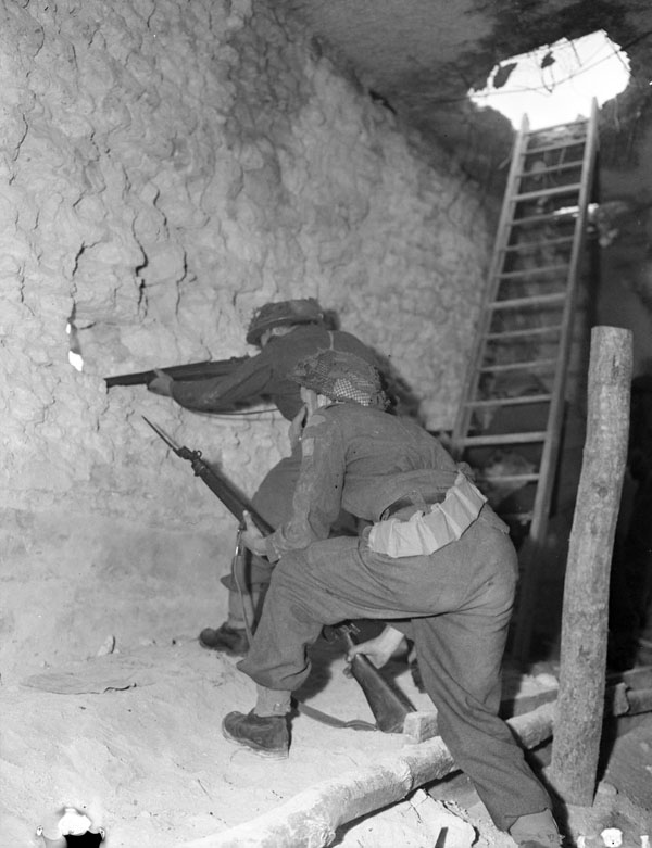 Riflemen D.H. Holmes and W.J. Wilkinson, both of The Regina Rifle Regiment, firing through loopholes in the wall of a captured barrack building, Vaucelles, France, 23 July 1943.