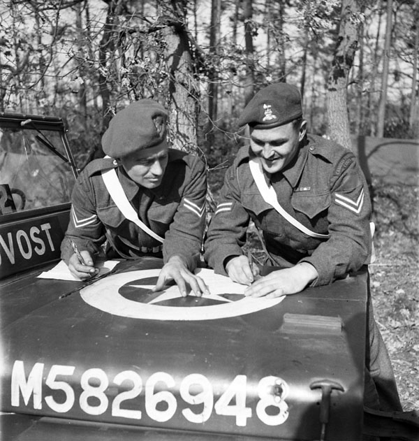 Two unidentified corporals of the 2nd Provost Company, Canadian Provost Corps (C.P.C.), exchanging notes on the hood of their jeep in the Reichswald, Germany, 20 March 1945.