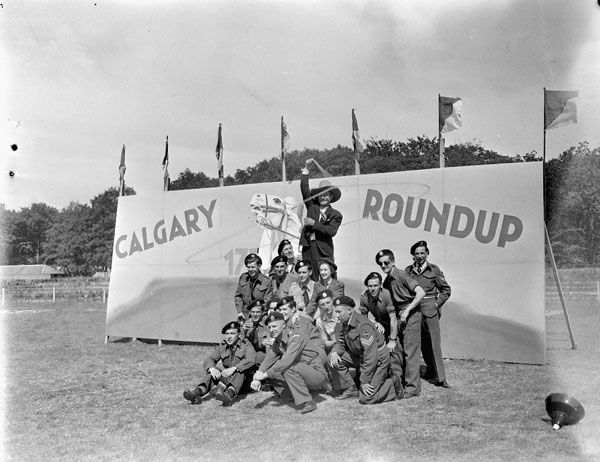 Entertainers and troopers of The Calgary Regiment in front of a sign reading “Calgary Roundup”, an event which included sports, entertainment, refreshments, and the presentation of a silver service to the regiment's commanding officer, Apeldoorn, Netherlands, 18 June 1945.