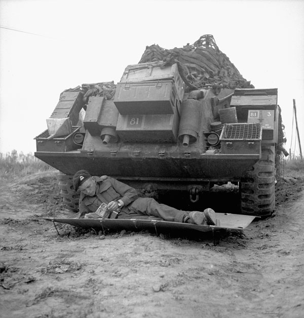 Trooper Ernie Tester of the 12th Manitoba Dragoons lying on a cot in front of a Sherman tank of a Canadian armoured regiment near Caen, France, 19 July 1944.