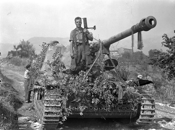 Lance-Corporal J.A. Thrasher of The Westminster Regiment (Motor), who holds the PIAT anti-tank weapon with which he disabled the German self-propelled 88mm. gun on which he is sitting, near Pontecorvo, Italy, 26 May 1944.