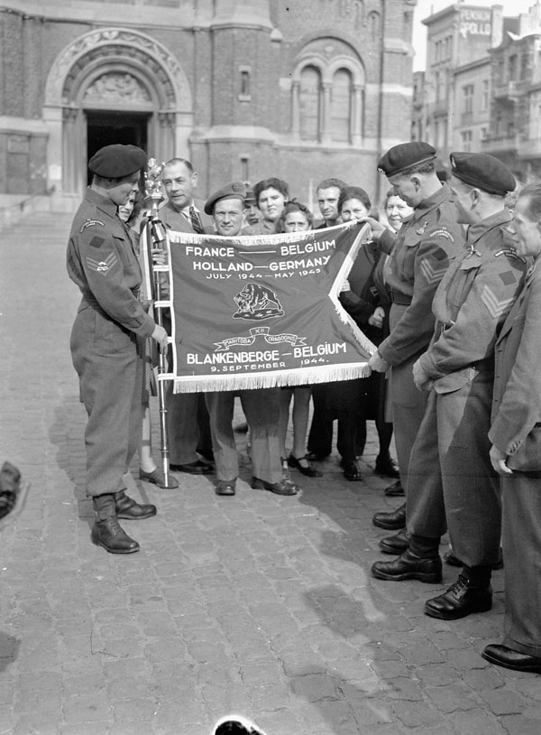 Personnel of the 12th Manitoba Dragoons holding a banner presented to the regiment by the town corporation of Blankenberghe, Belgium, 9 September 1945.