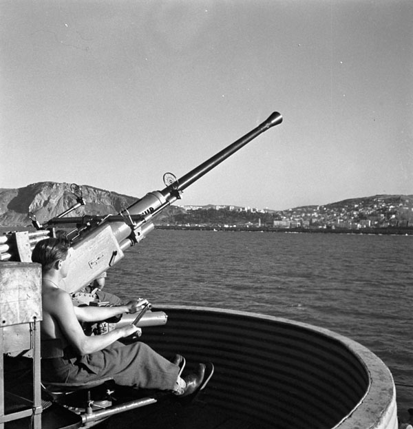 Private A.J. Landry manning a Bofors gun aboard the troopship H.M.T. NEA HELLAS, which is arriving at Philippeville, Algeria, 9 July 1943.